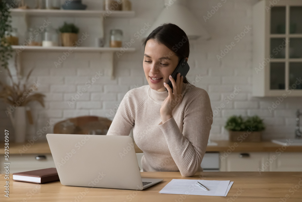 Positive engaged young freelance professional woman working in home kitchen, making mobile phone call, speaking to client, using online app on laptop computer, giving consultation, help, assist Positive engaged young freelance professional woman working in home kitchen, making mobile phone call, speaking to client, using online app on laptop computer, giving consultation, help, assist