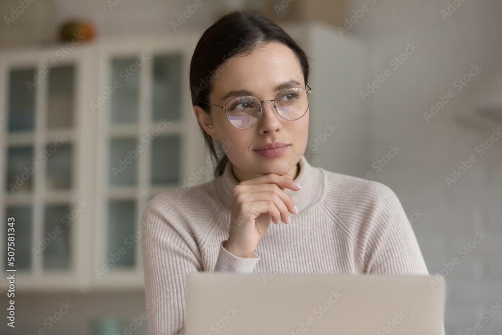 Thoughtful young laptop user in stylish eyeglasses sitting at home workplace in kitchen, looking away with pensive dreamy face, thinking of project future vision. Eyesight, eyeglasses wearing concept Thoughtful young laptop user in stylish eyeglasses sitting at home workplace in kitchen, looking away with pensive dreamy face, thinking of project future vision. Eyesight, eyeglasses wearing concept