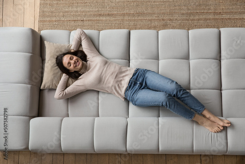 Relaxed cheerful beautiful 20s girl resting on soft comfortable big sofa, thinking, dreaming, smiling at good thoughts, planning future vacation, weekend, enjoying leisure at home, relaxing