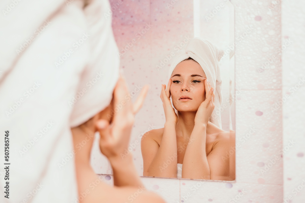 Young beautiful woman using a moisturizing facial mask after taking a bath. Pretty attractive girl in a towel on her head stands in front of a mirror in a home bathroom. Daily hygiene and skin care Young beautiful woman using a moisturizing facial mask after taking a bath. Pretty attractive girl in a towel on her head stands in front of a mirror in a home bathroom. Daily hygiene and skin care