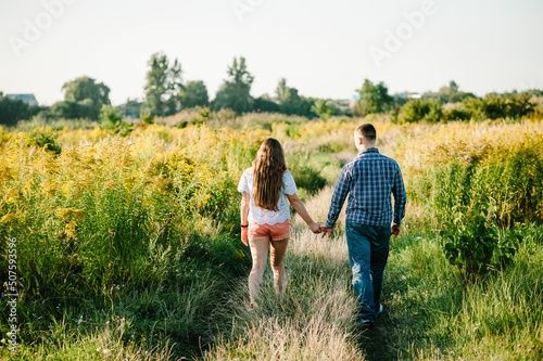 Rear view of a romantic man and woman walking on field grass, nature enjoying sunlight. Concept of lovely family holding hands. Young couple running and looking away. Back view.