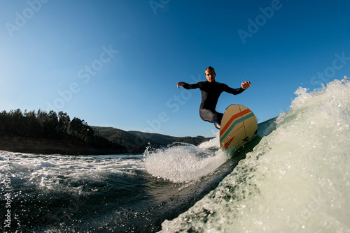 man wakesurfer riding down the splashing wave on blue sky background
