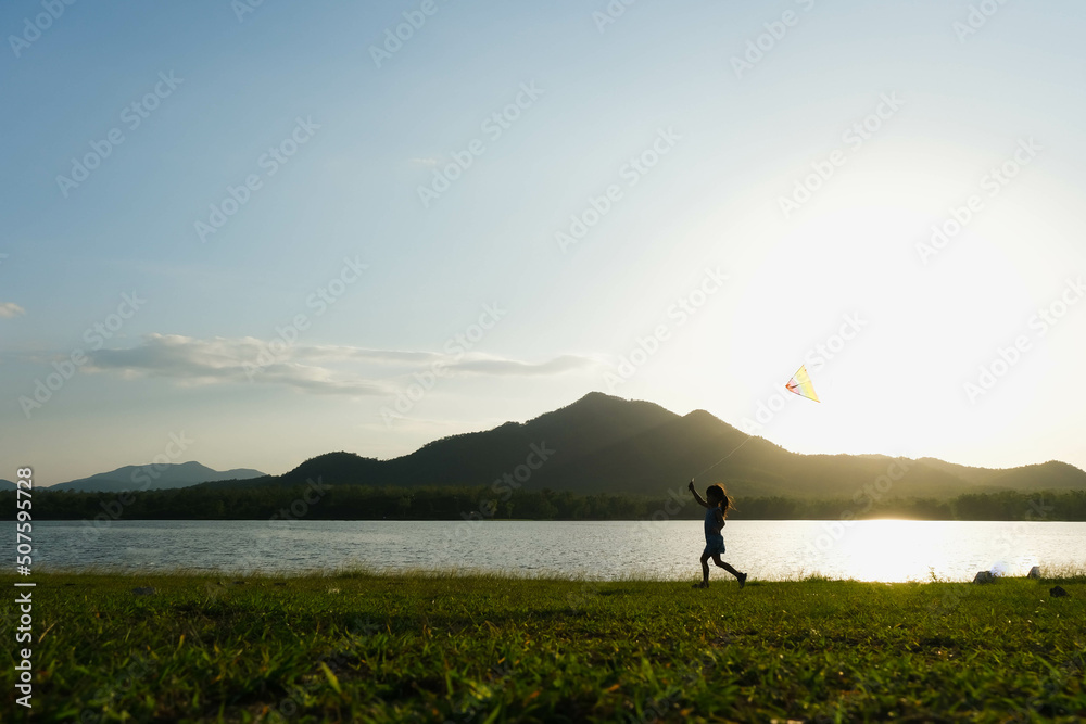 Child playing with a kite while running on a meadow by the lake at sunset. Healthy summer activity for children. Funny time with family.