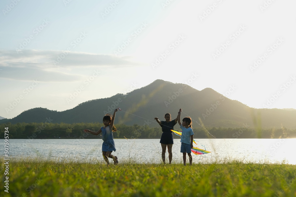 Children are flying kites while running on a meadow by the lake at sunset with their mother. Healthy summer activity for children. Funny time with family.