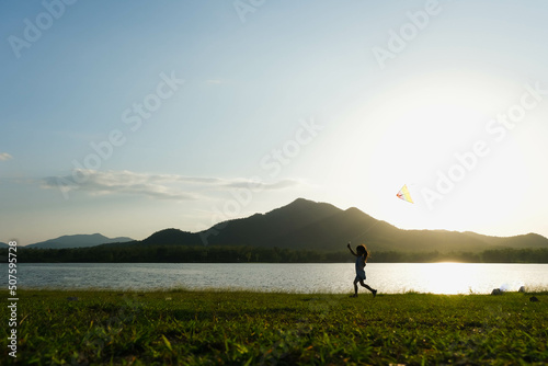 Fototapeta Naklejka Na Ścianę i Meble -  Child playing with a kite while running on a meadow by the lake at sunset. Healthy summer activity for children. Funny time with family.