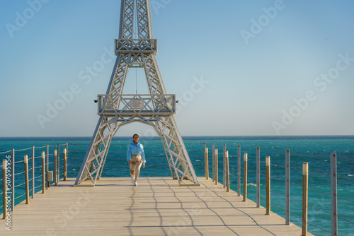 Large model of the Eiffel Tower on the beach. A woman walks along the pier towards the tower  wearing a blue jacket and white jeans.