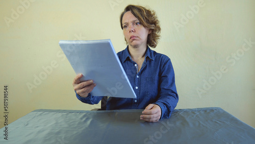 A forty-year-old woman, blond in a blue shirt, looks at a white paper while sitting at a table.