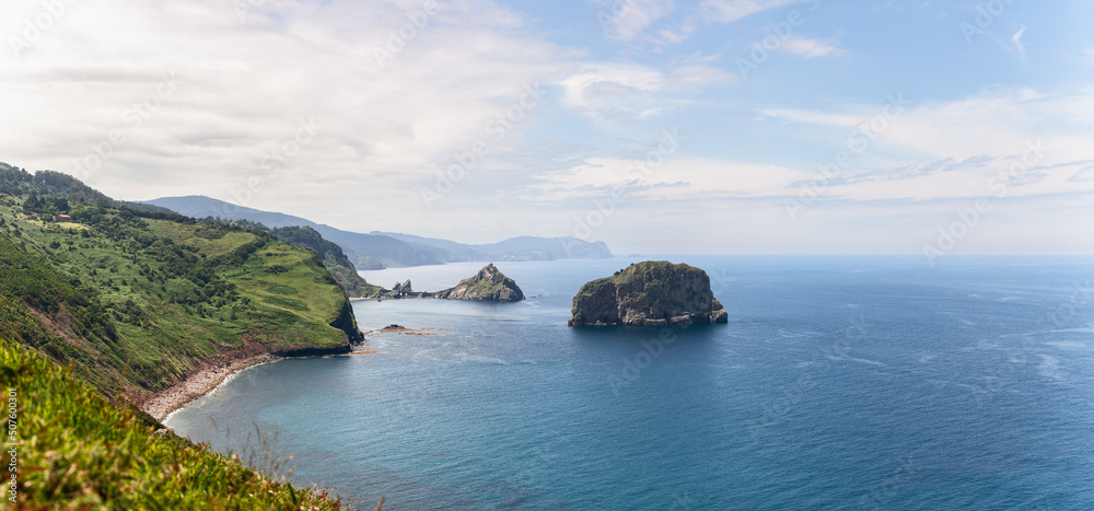 Wide panorama of emerald green cape cabo Machichaco of Basque coast and two small islands Gaztelugatxe and Aketx. Cantabrian Sea, stretching beyond the horizon, Spain