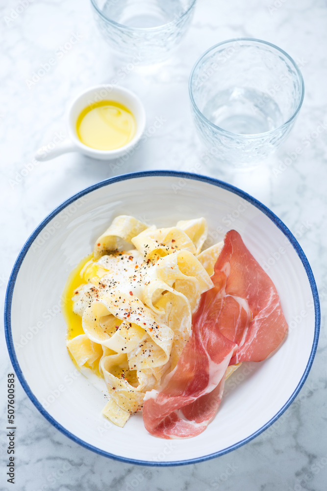 Creamy pappardelle with prosciutto and poached egg in a white plate, vertical shot on a light-grey marble background Creamy pappardelle with prosciutto and poached egg in a white plate, vertical shot on a light-grey marble background