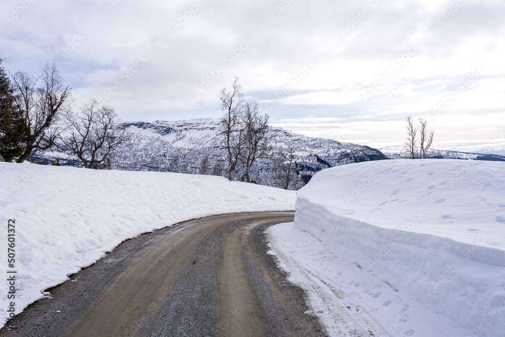 Dirt road in the mountains of Norway in winter when a lot of white snow has reached along the edges and there are white clouds behind the sun shining in the blue sky Dirt road in the mountains of Norway in winter when a lot of white snow has reached along the edges and there are white clouds behind the sun shining in the blue sky