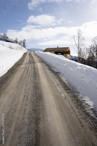 Dirt road in the mountains of Norway in winter when a lot of white snow has reached along the edges and there are white clouds behind the sun shining in the blue sky