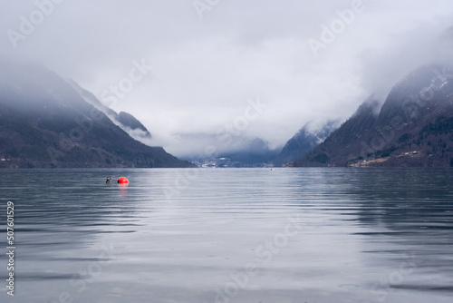 Norwegian fjord with clear water in early spring when there is still some white snow in the mountains and they are foggy, but in the dark waters of the fjord you can see a bright red buoy