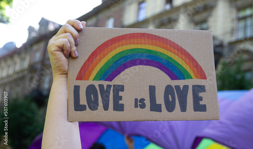 Woman holding placard sign Love is Love with rainbow, symbol of LGBT. Giant flag in background. Pride Parade, equality march to support and celebrate LGBT+, LGBTQ gay and lesbian community. © Longfin Media