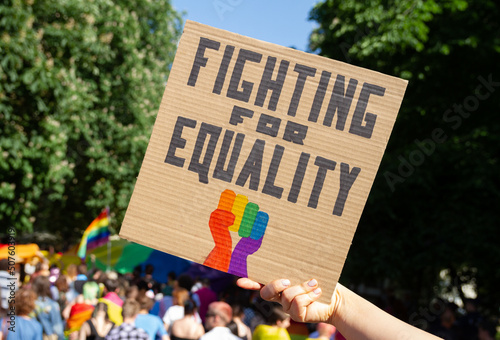 Woman holding placard sign Fighting for Equality with rainbow flag fist, during LGBT Pride Parade. Crowd of people at equality march to support and celebrate LGBT+, LGBTQ gay and lesbian community.