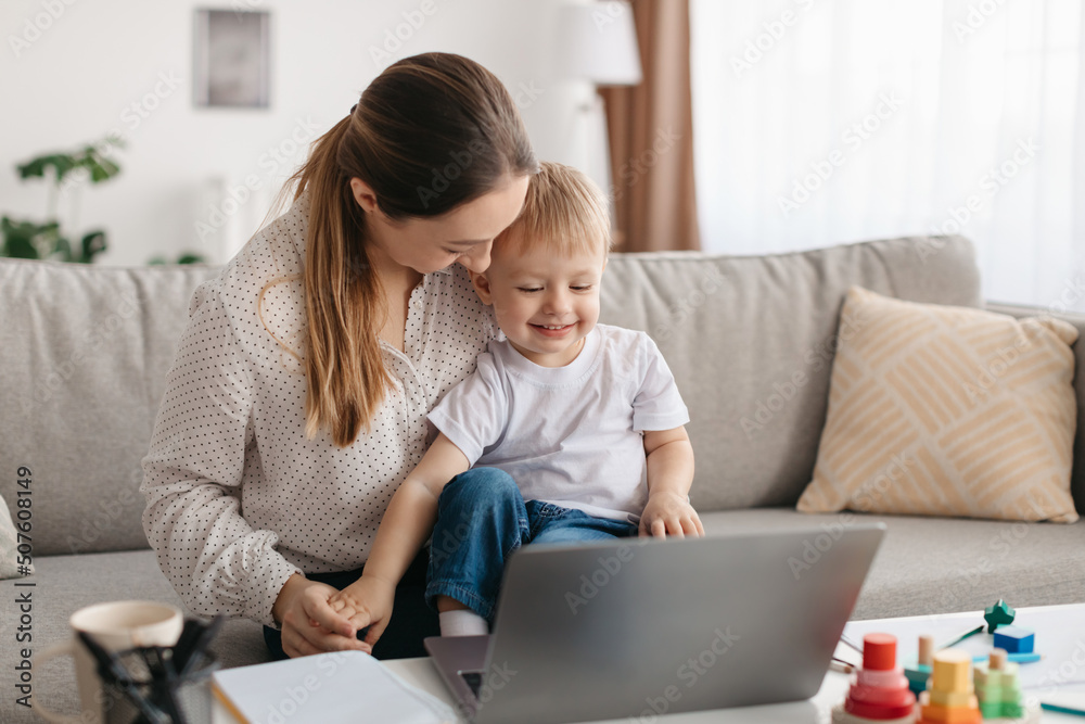Loving young mom playing with son while working on laptop, sitting on sofa and tickling child, enjoying maternity leave Loving young mom playing with son while working on laptop, sitting on sofa and tickling child, enjoying maternity leave