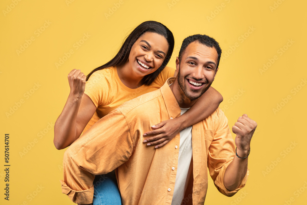 Obraz premium Attractive young African American man giving piggyback ride to his girlfriend, gesturing YES on yellow studio background