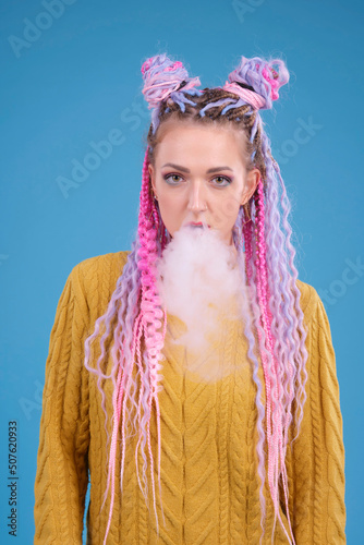 Young woman with colored colorful hair smoking electronic cigarette, blowing smoke from mouth, studio portrait. Bright woman with afrobraids. Blue solid background. Arrogant person photo