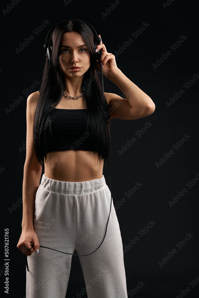 Gorgeous dark haired wonan in sporty outfit posing with earphones on head indoor. Front view of serious lady looking at camera and standing straight, isolated on black background. Concept of posing. Gorgeous dark haired wonan in sporty outfit posing with earphones on head indoor. Front view of serious lady looking at camera and standing straight, isolated on black background. Concept of posing.