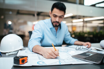 Concentrated serious attractive millennial arab man engineer with beard works with drawings at workplace