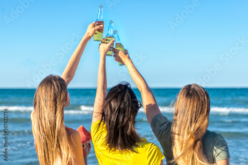 three happy girls toasting beers looking at the horizon beach outdoors celebrating summer vacation days. faceless friends having fun on seaside holidays at sunset. joy, freedom, genz and party concept © blackandbrightph