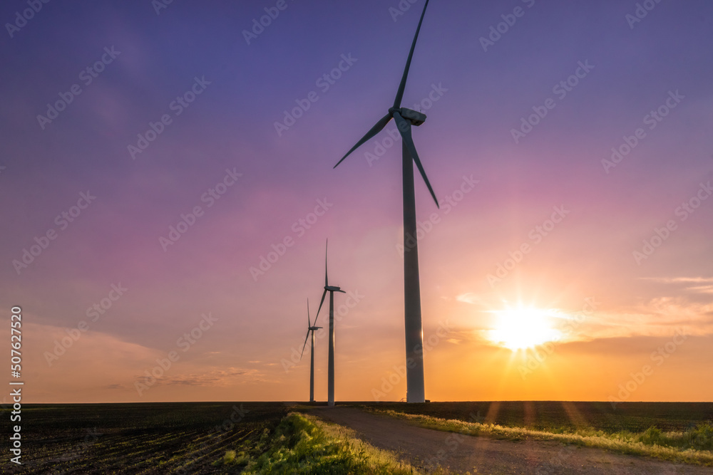 Obraz premium Two windmills standing tall in a mid-western field during a vibrant, colorful sunset. 