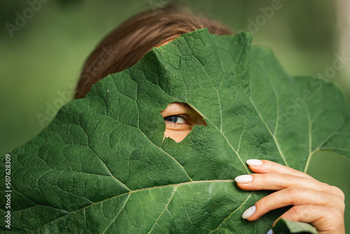 Canvastavla a young girl looks into the camera through a hole in a large burdock leaf