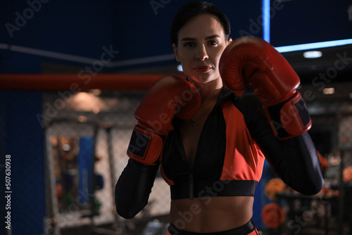 Female in red boxing gloves and sportive wear stands in a rack in the boxing hall. © ty