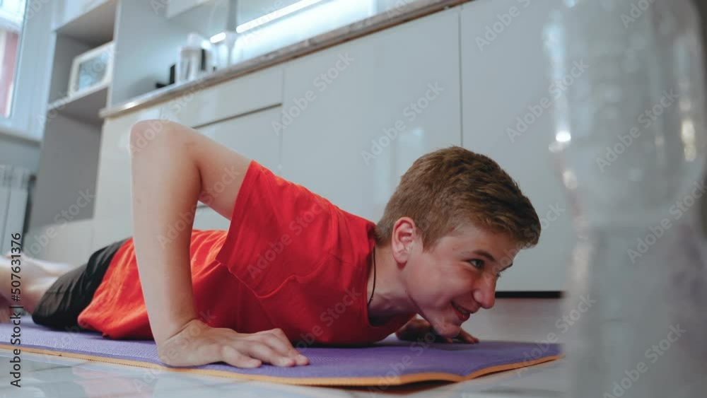 Skinny teen boy goes in for sports in quarantine in kitchen on yoga mat. Guy does push-up exercise through strength and lies on floor from exhaustion but is satisfied. Male smiles Skinny teen boy goes in for sports in quarantine in kitchen on yoga mat. Guy does push-up exercise through strength and lies on floor from exhaustion but is satisfied. Male smiles