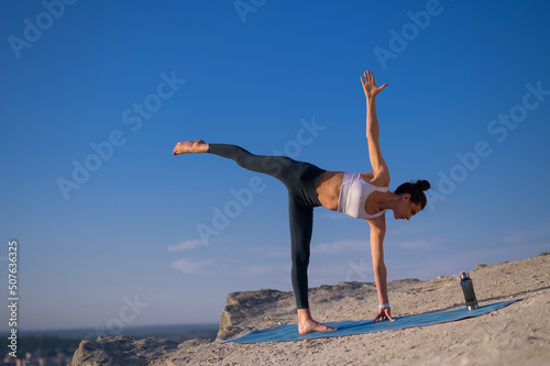 Beautiful slender caucasian girl in sportswear practices yoga on a rock on a background of blue sky at sunset