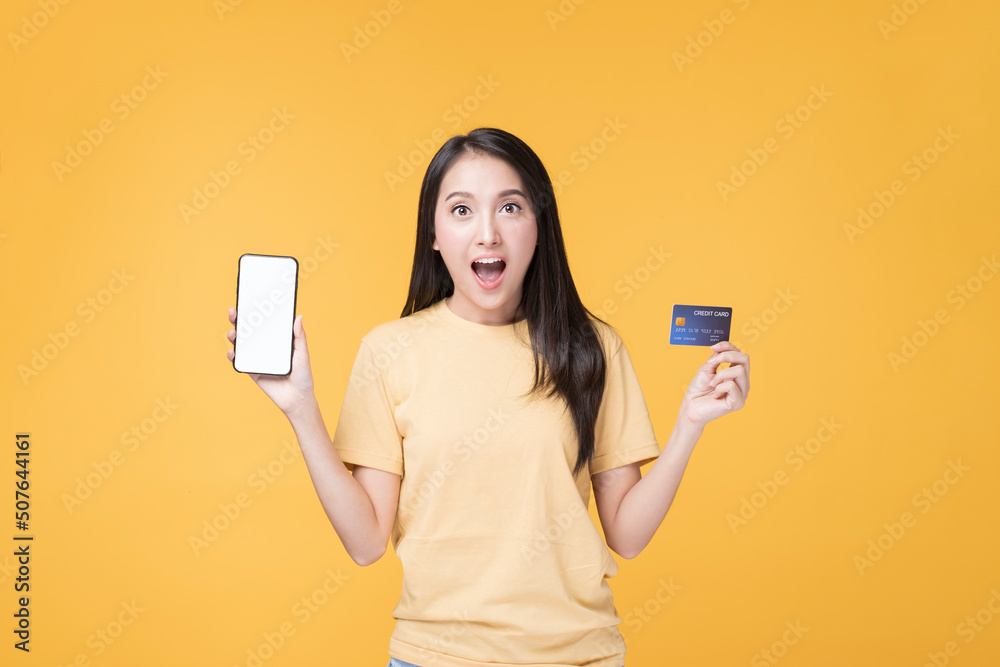 Portrait of Happy Asian female holding Credit Card. Young Asian Woman holding Credit card and Smartphone. Happy Asian Woman wearing Yellow T-shirt standing over yellow background. Portrait of Happy Asian female holding Credit Card. Young Asian Woman holding Credit card and Smartphone. Happy Asian Woman wearing Yellow T-shirt standing over yellow background.