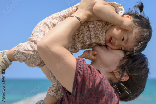 【沖縄・画像】青空と青い海を背景にお母さんが娘を抱きかかえて遊んでいる写真