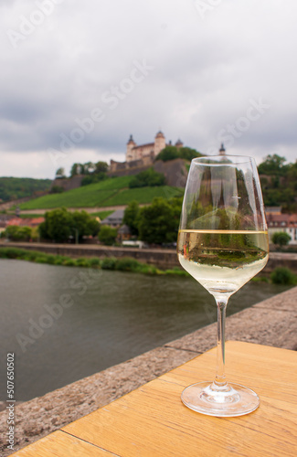 A glass of white wine standing on a wine table on an Old Bridge on Main river in front of Marienberg Fortress upon a vineyard hills in Bavarian touristic city