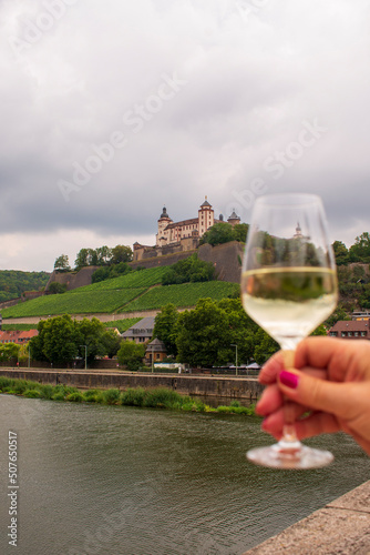 Woman hand holding a glass of white wine on an Old Bridge on Main river in front of Marienberg Fortress upon a vineyard hills in Bavarian touristic city  