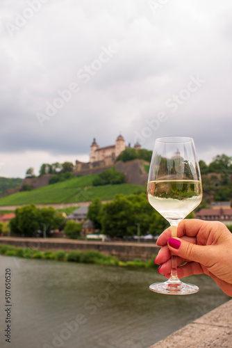 Woman hand holding a glass of white wine on an Old Bridge on Main river in front of Marienberg Fortress upon a vineyard hills in Bavarian touristic city  