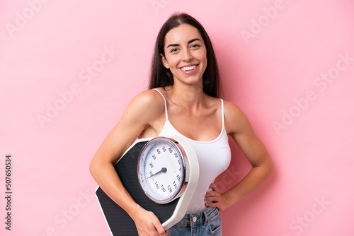 Young caucasian woman isolated on pink background with arms at hip and holding weighing machine