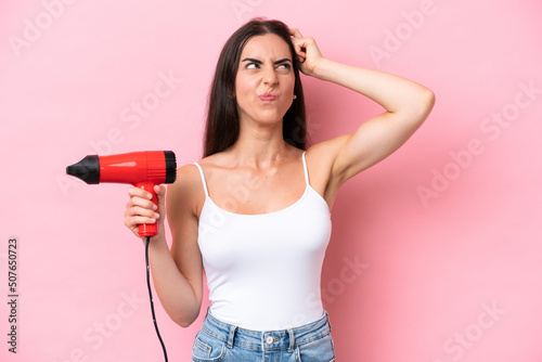 Young caucasian woman holding a hairdryer isolated on pink background having doubts and with confuse face expression © luismolinero