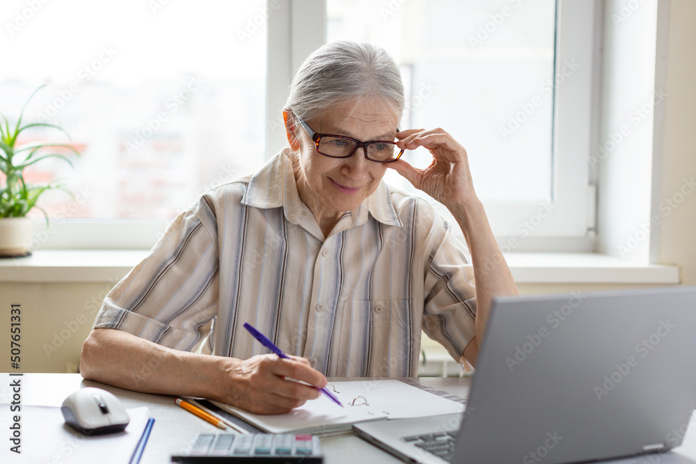 Intelligent caucasian senior woman adjust her glasses and look at laptops screen with a pen in hand, indoor, selective focus. Intelligent caucasian senior woman adjust her glasses and look at laptops screen with a pen in hand, indoor, selective focus.