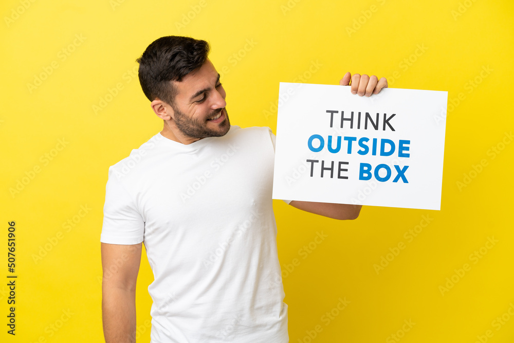 Young handsome caucasian man isolated on yellow background holding a placard with text Think Outside The Box Young handsome caucasian man isolated on yellow background holding a placard with text Think Outside The Box