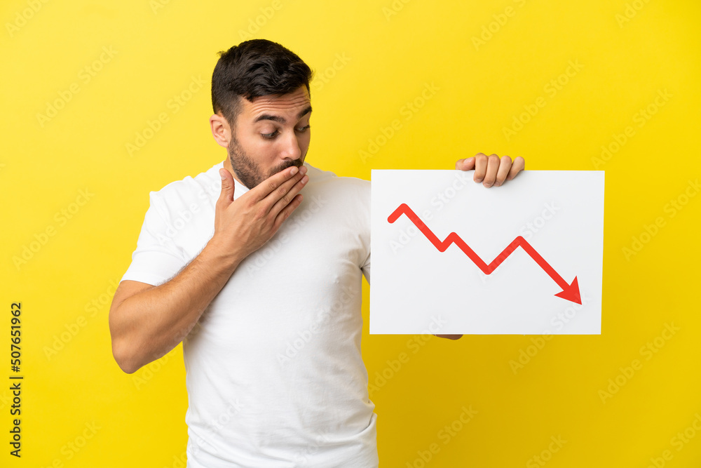 Young handsome caucasian man isolated on yellow background holding a sign with a decreasing statistics arrow symbol with surprised expression Young handsome caucasian man isolated on yellow background holding a sign with a decreasing statistics arrow symbol with surprised expression