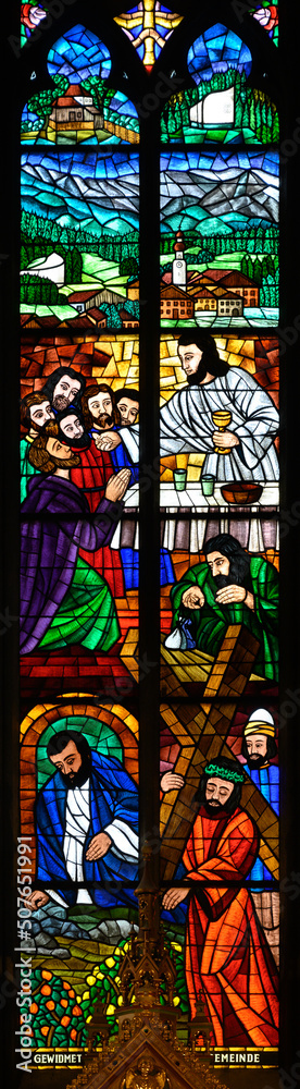 Stained-glass window depicting the Last Supper and Jesus carrying the cross. Above is the town of Erl in Tyrol with its church. Votivkirche – Votive Church, Vienna, Austria. 2020-07-29.