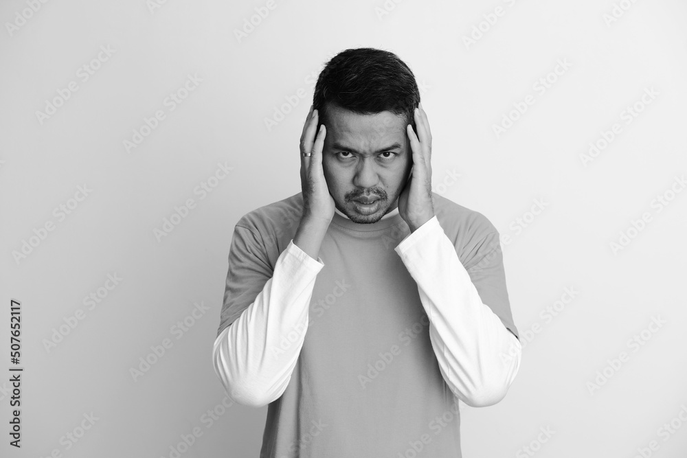 Adult Asian man looking to camera with both hand touching his head showing depression Adult Asian man looking to camera with both hand touching his head showing depression