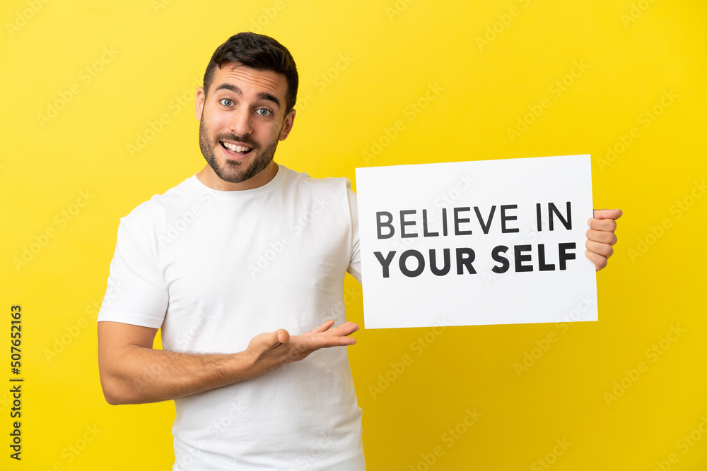 Young handsome caucasian man isolated on yellow background holding a placard with text Believe In Your Self and pointing it Young handsome caucasian man isolated on yellow background holding a placard with text Believe In Your Self and pointing it