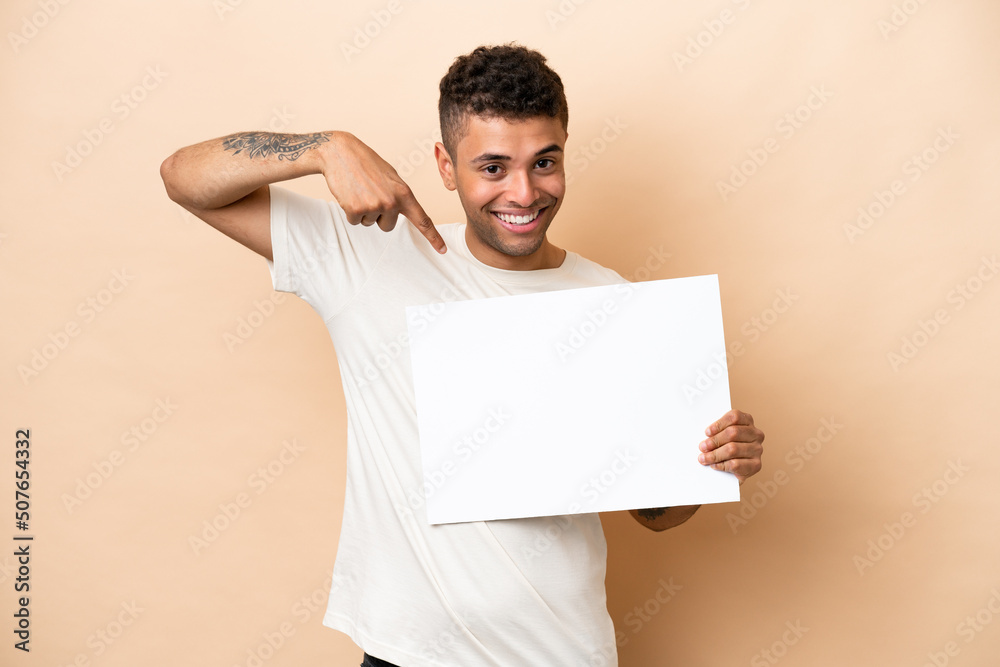Young Brazilian man isolated on beige background holding an empty placard with happy expression and pointing it Young Brazilian man isolated on beige background holding an empty placard with happy expression and pointing it