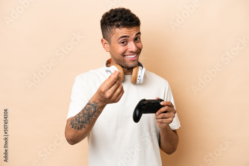 Young Brazilian man playing with a video game controller isolated on beige background making money gesture