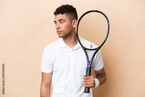 Young brazilian handsome man playing tennis isolated on beige background looking to the side © luismolinero