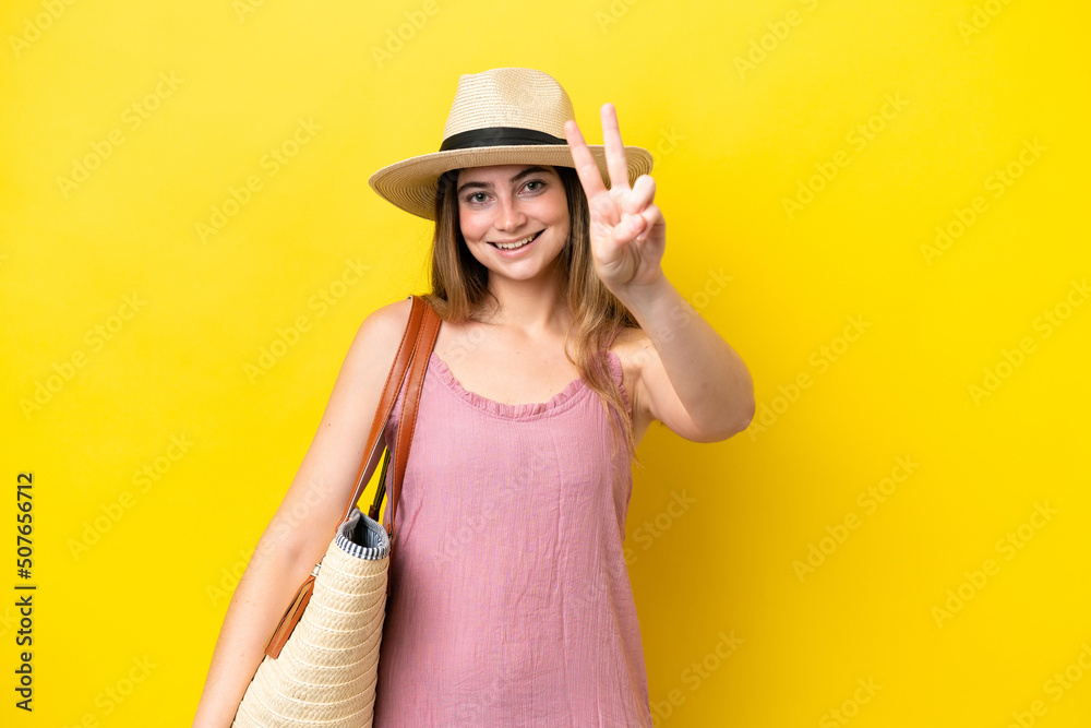 Young caucasian woman holding a beach bag isolated on yellow background smiling and showing victory sign Young caucasian woman holding a beach bag isolated on yellow background smiling and showing victory sign