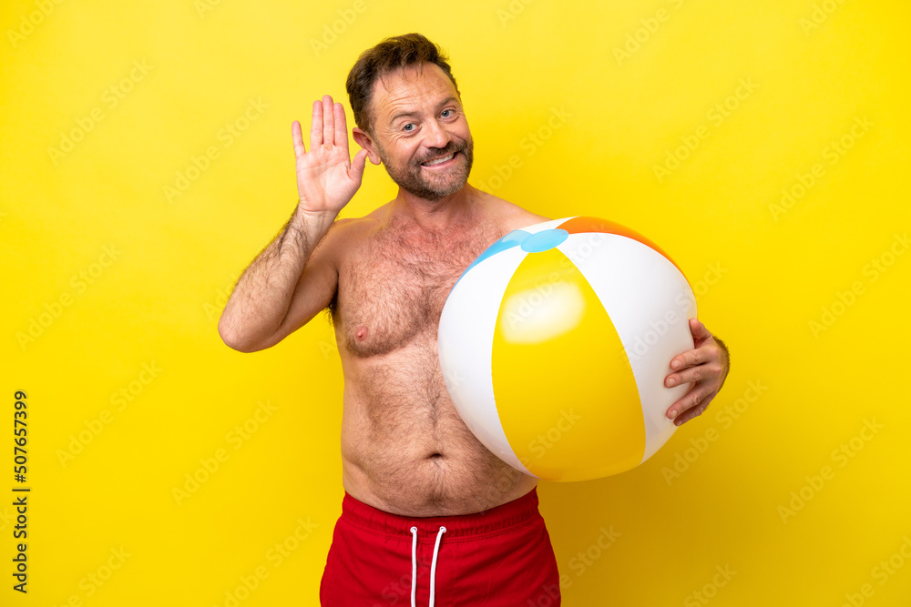 Middle age caucasian man holding beach ball isolated on yellow background saluting with hand with happy expression