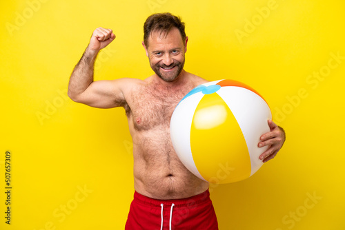 Middle age caucasian man holding beach ball isolated on yellow background doing strong gesture