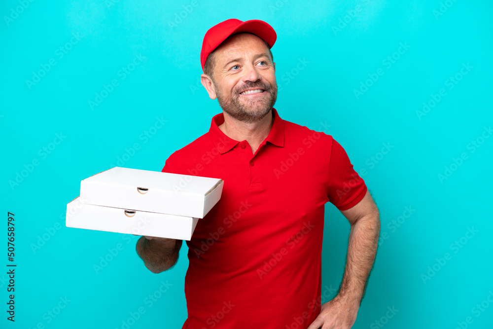 Pizza delivery man with work uniform picking up pizza boxes isolated on blue background posing with arms at hip and smiling