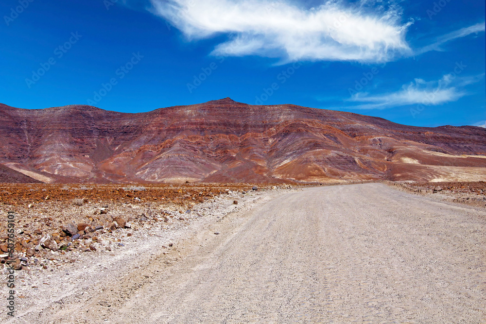 Dirt road in arid barren dry landscape, red mountains, clear blue sky - Road trip to Punta de Jandia, Fuerteventura